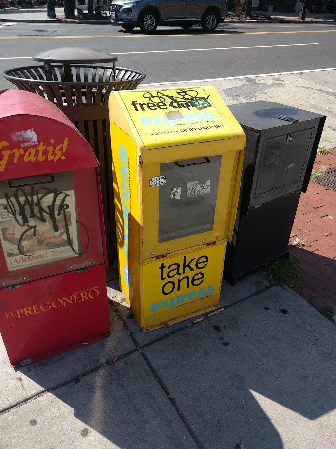 An yellow Express daily newspaper box with no papers sits between two other distribution boxes on a street corner in Washington, DC. A photograph by Michael Lee from September 19, 2019.