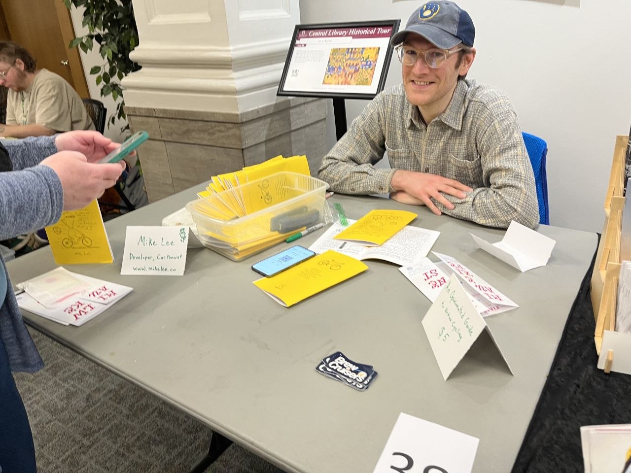 A photograph of me vending a zine fest. A zine with a yellow cover on display on a foldable table. Zines stack in a plastic container.